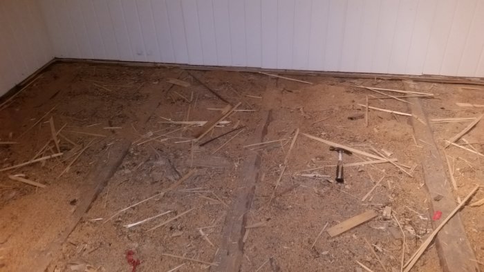 Oorganiserad hög av träavfall och spikar på ett betonggolv med vit panelvägg i bakgrunden.