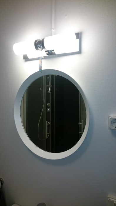 Oval spegel på vägg med reflektion av duschkabin, över spegeln en vägglampa med två ljuskällor.