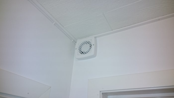 Väggventilator i ett hörn av ett nyrenoverat badrum monterad på en vit texturerad vägg ovanför taklist.