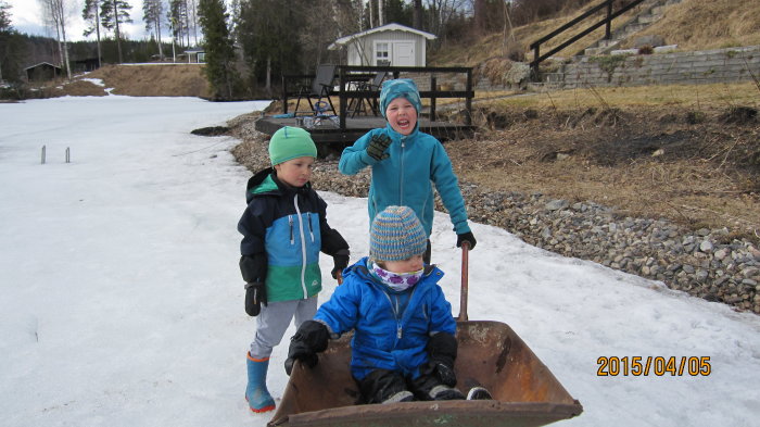 Tre barn leker nära en grushög på en isbelagd yta, ett barn sitter i en rostig skottkärra.
