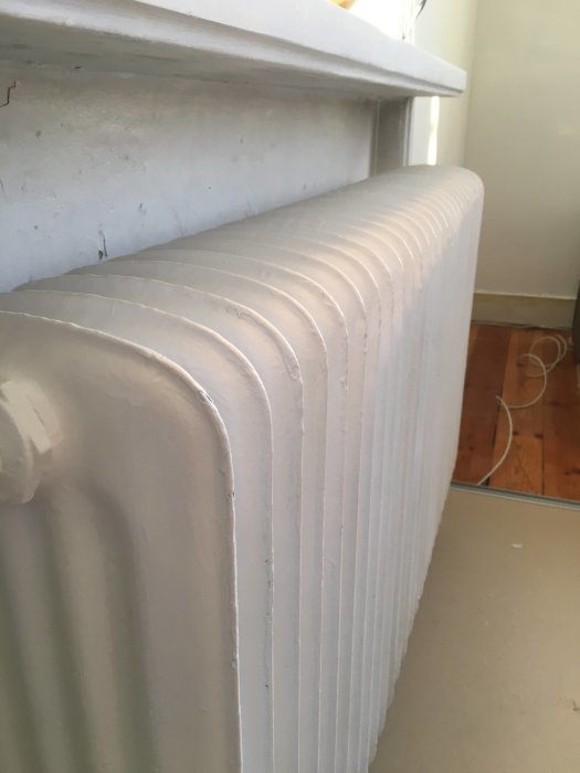 Nymålat radiator i grå-beige nyans, sprayfärgad för en jämn yta, med en del av fönsterkarmen synlig.