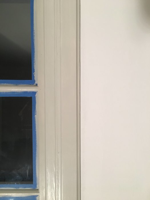 Nymålad dörrkarm i grå-beige färg bredvid ett fönster med tejpade kanter i förväntan på målning.