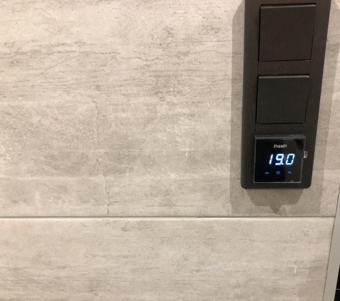 Väggmonterad termostat visar 19.0 grader, bredvid strömbrytare, på en texturerad ljus vägg.
