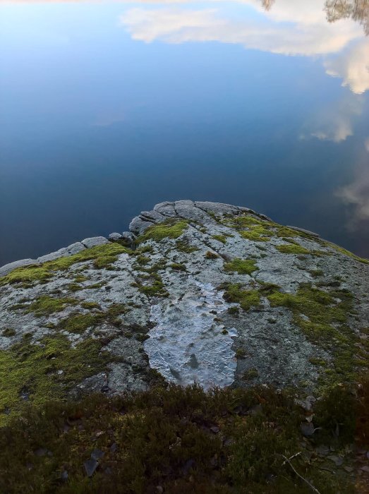Stenig kust med mossbeväxta stenar vid lugnt vatten som speglar himlen.