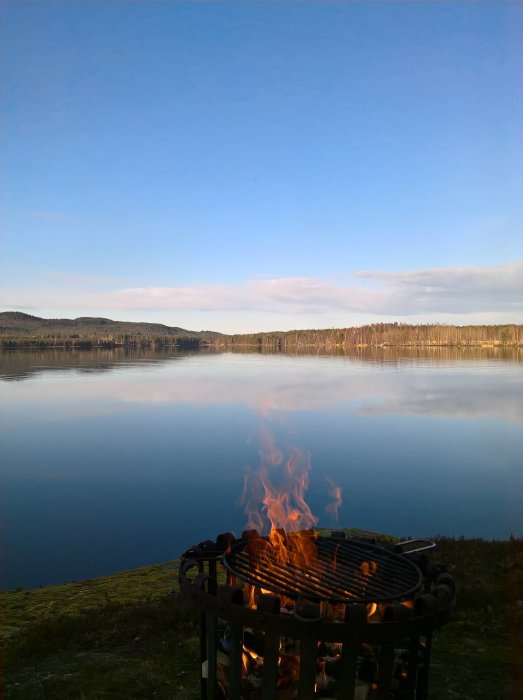 Grill med tänd eld vid en stilla sjö med spegelblank vattenyta och skog i bakgrunden.