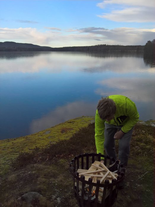 Person förbereder eld i korg för grillning vid sjö med lugnt vatten och skog i bakgrunden.