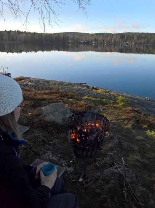 Person sitter vid tänd grillkorg nära en stilla sjö en kylig dag.