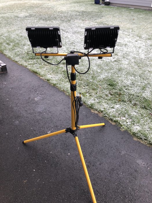 Två arbetslampor monterade på ett gult stativ på en asfalterad yta med lätt snötäcke i bakgrunden.