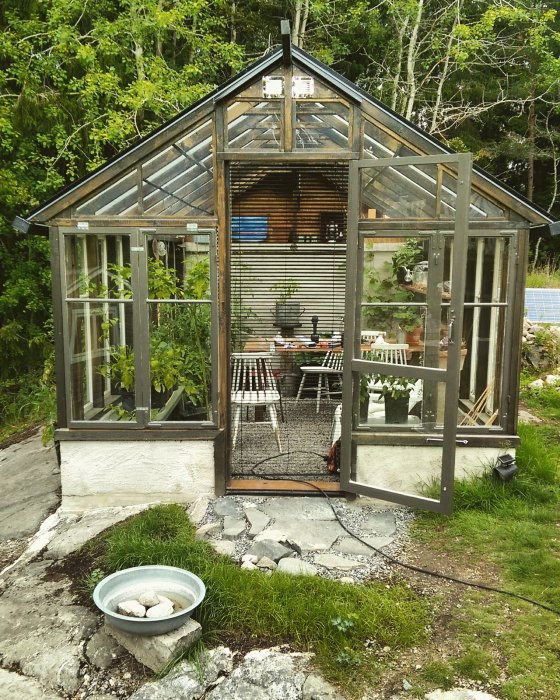 Växthus av glas och metall med öppen dörr, visar inredning med bord och planteringar, omgivet av grönska.