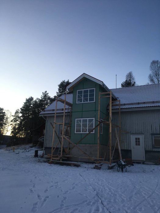 Hus under renovering med tegel och hängrännor i vintersnö, medan byggställningar och bar mark syns i förgrunden.