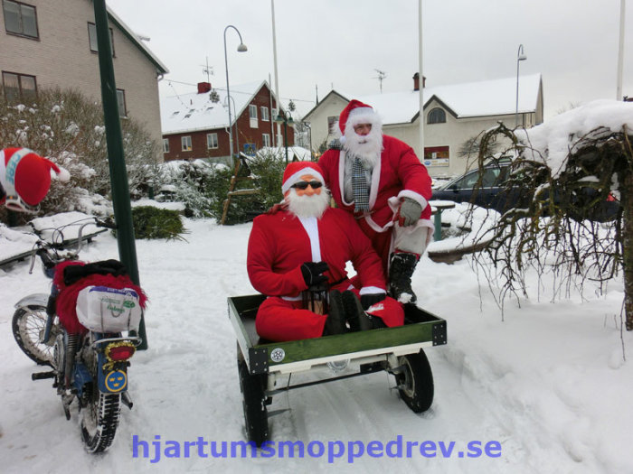 Två personer utklädda till tomtar sitter på en moped med släp i ett vinterlandskap.