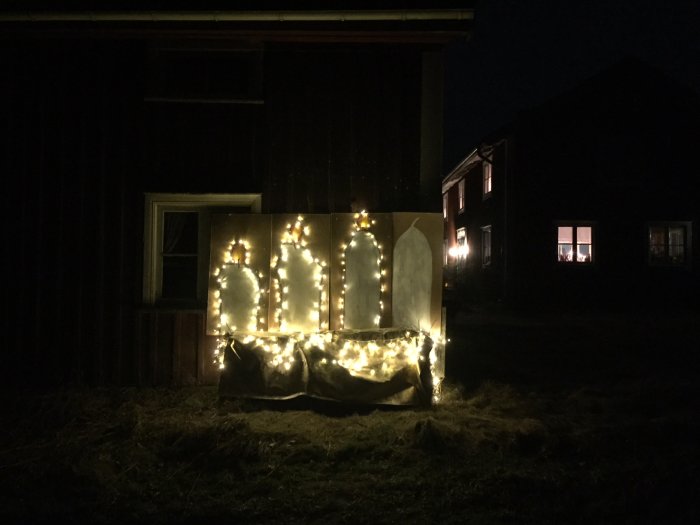 Juldekoration med fyra hemmagjorda ljusstakar belysta av slingor placerade framför en mörk byggnad på kvällen.