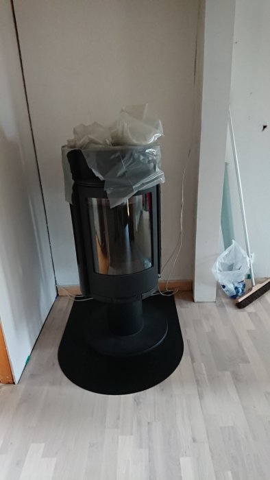 Modern kamin insvept i plast står på en svart platta i ett rum med laminatgolv, redo för användning.