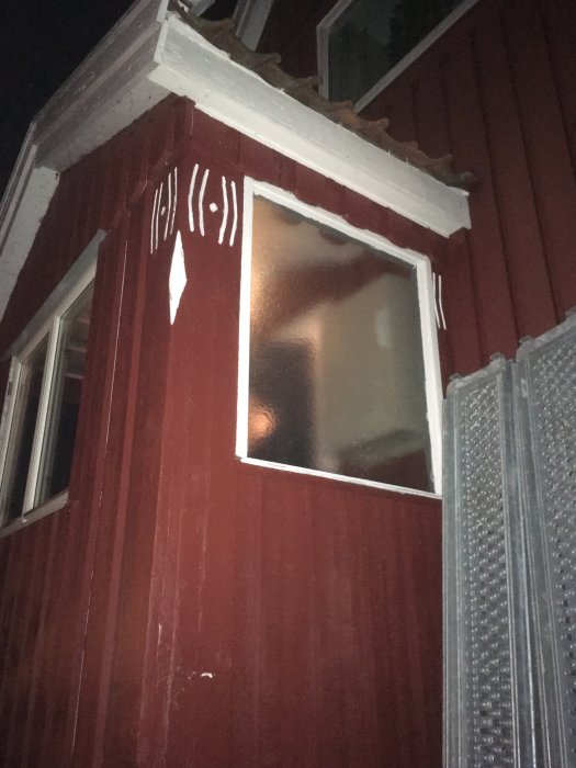 Utskuren signatur eller markering på stolpe av ett rött hus med vitmålade kanter och ett fönster, tagen på kvällen.