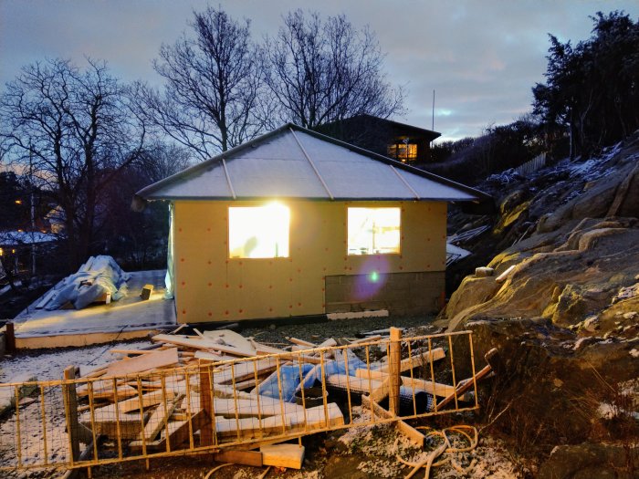 Nybygge av hus i skymningen med belysning inifrån och byggmaterial framför.