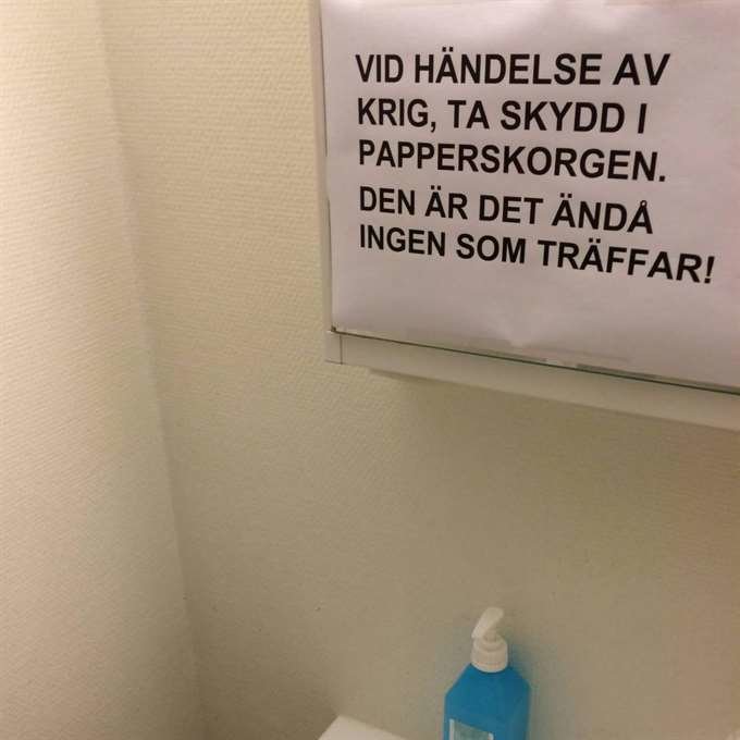 Vägg med skämtsamt textat papper och handspritflaska på bänk.