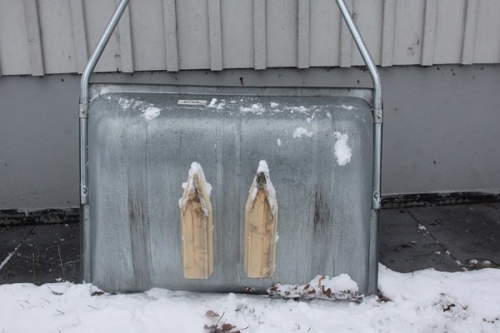 Spårmaskin för skidspår med snötäckta trätrissor framför en grå vägg.