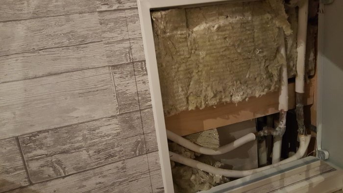 Öppnad vägg med synlig isolering och rörledningar bakom träpanel.