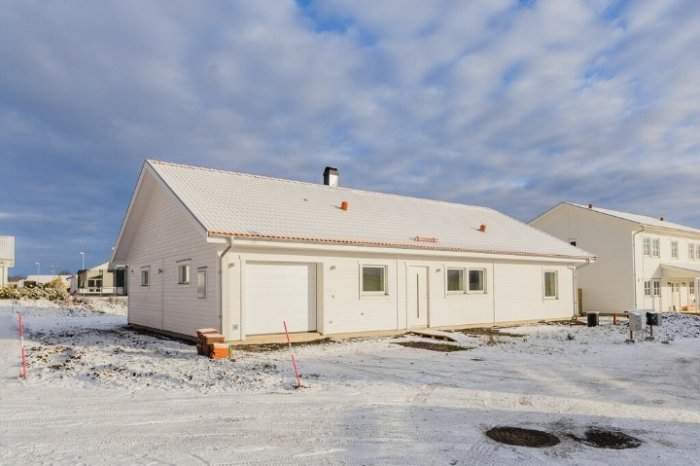 Nybyggt vitt enplanshus med garage under lätt snötäcke mot blå himmel.