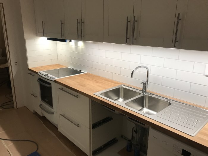 Kök under renovering med vita skåp, träbänkskiva, dubbelho och inbyggd diskmaskin.