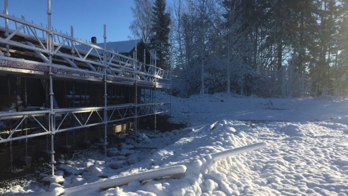 Hus under konstruktion med byggnadsställningar, täckt av snö i soligt vinterlandskap.