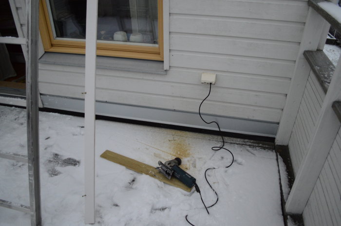 Bild av balkong med tätad springa nära masonitvägg, caulk gun och bräda på snöig mark.