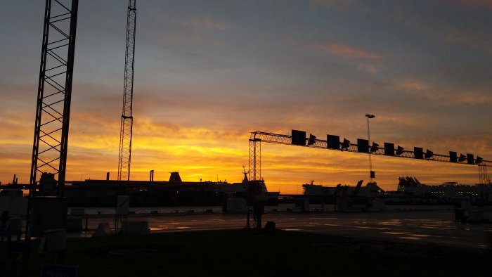 Soluppgång över hamnen med silhuetter av kranar och trafikljus mot en färgrik himmel.