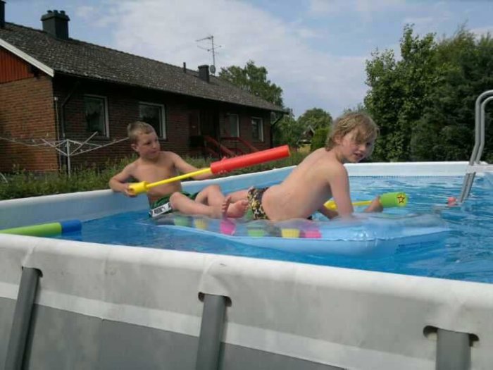 Två barn leker i en ovanmarkspool med leksaker och flythjälpmedel, bakom syns ett rött hus.