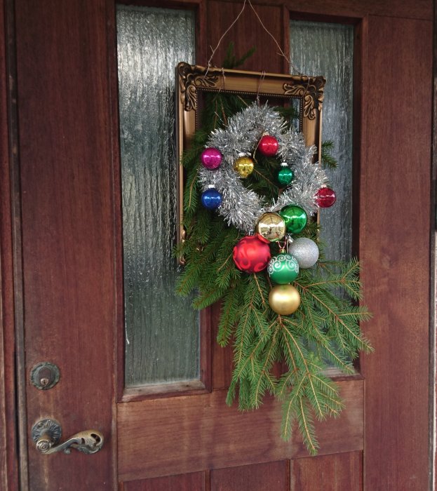 En julkrans med färgglada kulor och silverglitter hängande på en brun dörr med frostigt glas.