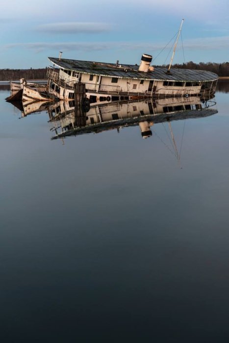 Ett sjunkande skepp i stilla vatten med speglingar, omgivet av himmel och hav.