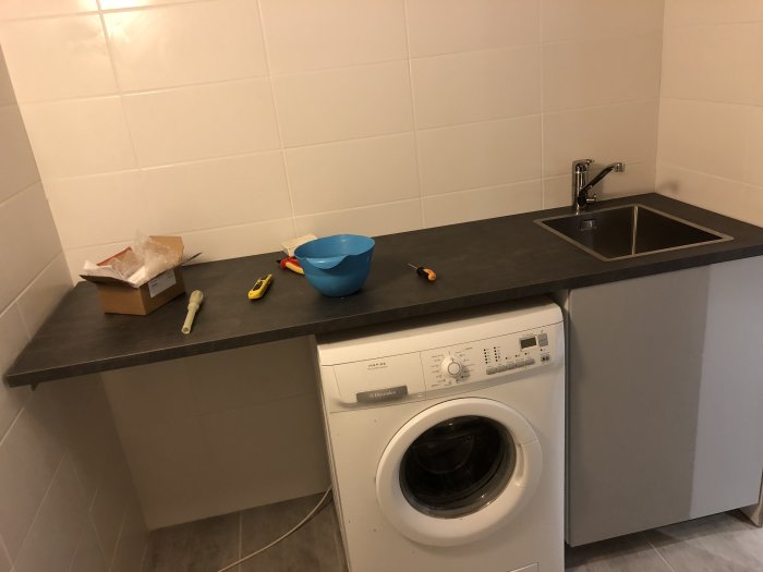 Tvättmaskin under arbetsbänk med diskho, verktyg och skålar på bänken i ett kaklat rum.