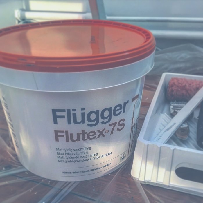 Ett öppet hink med Flügger Flutex 7S väggfärg och en färgroller i en färgkaret på en presenning.