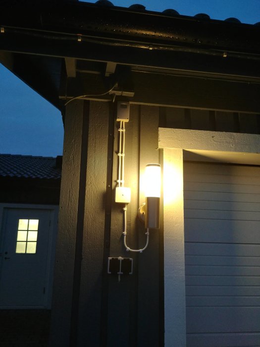 Utomhusvägglampa med synlig kabeldragning från kopplingsdosa vid husfasad i skymningen.