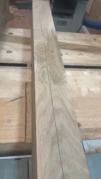 Stor träplanka med kvistar och längsgående djupa sprickor ligger på en arbetsbänk i en verkstad.