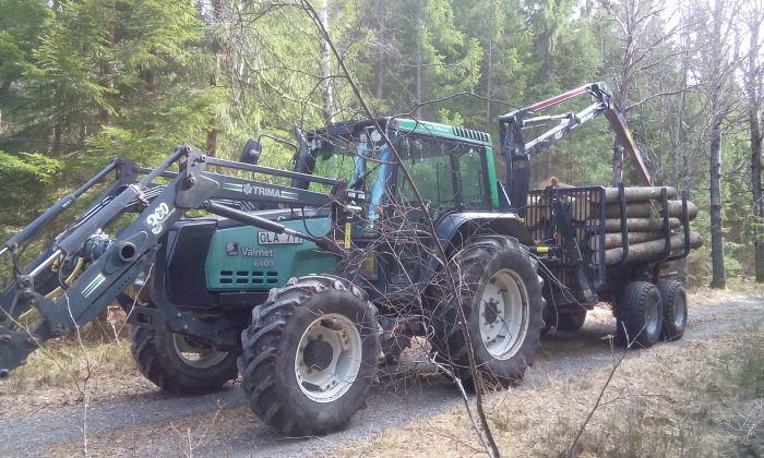 Grön Valmet 6400 traktor med skogsvagn fullastad med timmer i skogsmiljö.