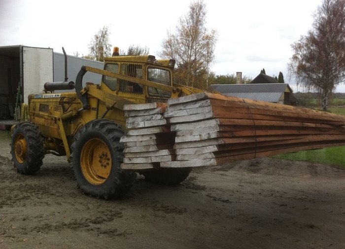 Gul Volvo BM 641 baklastare med fyrhjulsdrift lyfter virkeshögar utanför en byggnad.