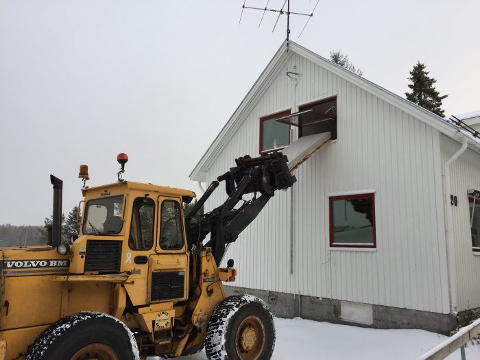 Gul Volvo BM baklastare framför ett vitt hus i vintermiljö, snö på marken och utrustad med lyftarm.