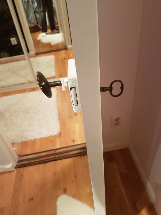 Gammaldags dörrhandtag och nyckel i en vit innerdörr, med utsikt till trägolv och karm.
