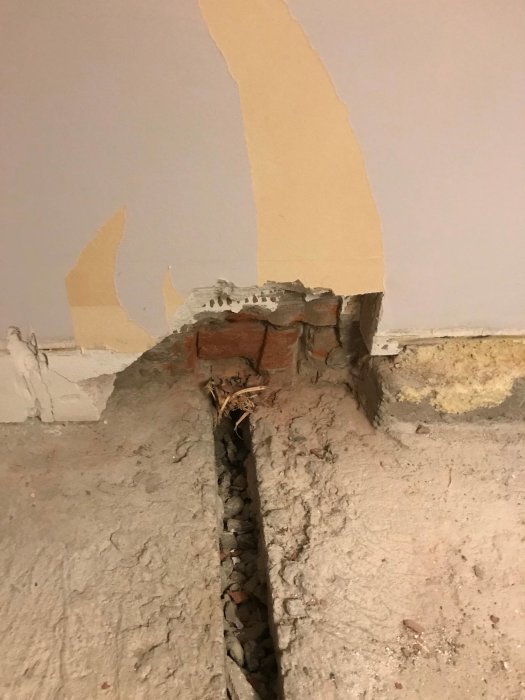 Skadad vägg och golv med en öppen ränna avbeton och exponerade tegelstenar som behöver repareras.