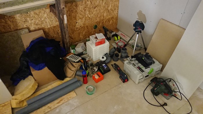Hörn av ett byggarbetsrum med diverse verktyg, byggmaterial och VVS-utrustning utspridda på golvet.