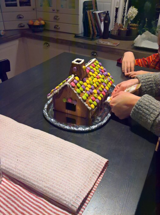 Ett pepparkakshus dekorerat med färgglada godis på ett bord, händer som dekorerar med smält choklad.