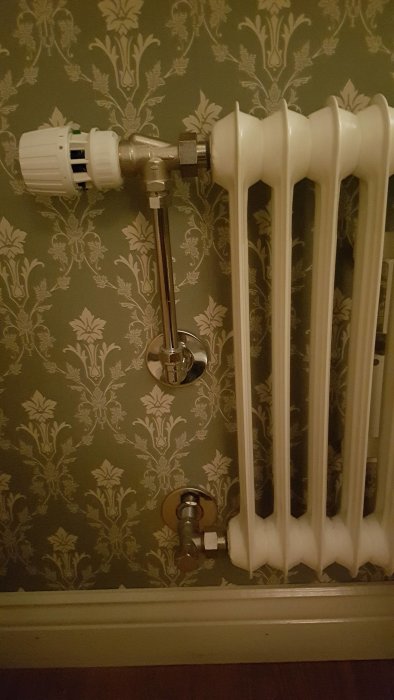 Vit elementradiator installerad mot en vägg med mönstrad tapet, komplett med termostat och rörkopplingar.