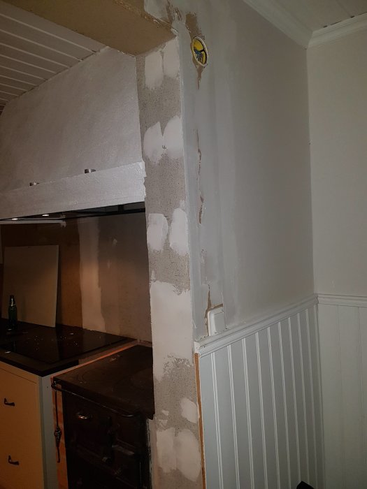 Ofärdig köksrenovering med naken puts, spiskåpa och vedspis intill vit bröstpanel.