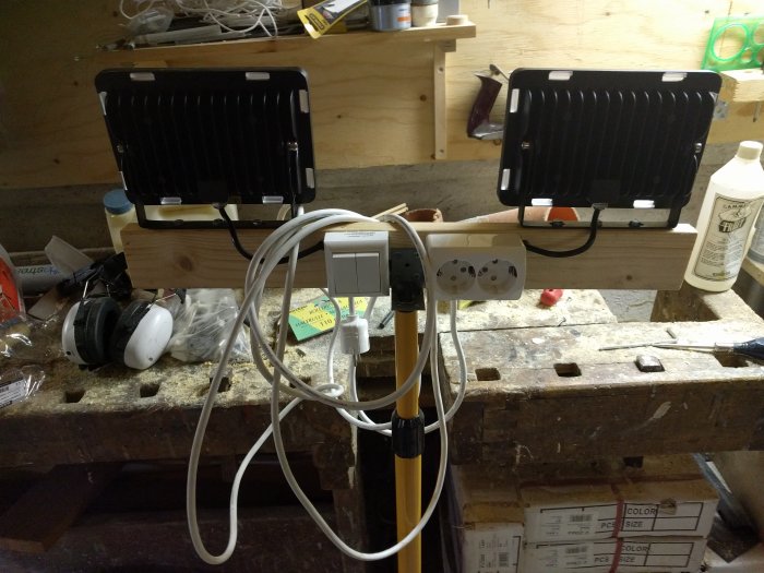 Två LED-arbetslampor monterade på en träregel ovanför en verktygsbänk med diverse verktyg och material.