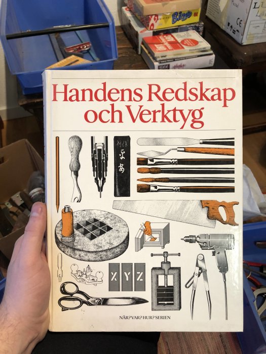 Hand håller en bok om verktyg med titeln "Handens Redskap och Verktyg