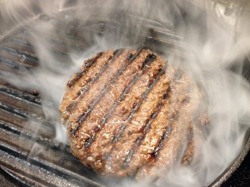 Hemgjord högrevsburgare på grillpanna med rök som stiger upp.