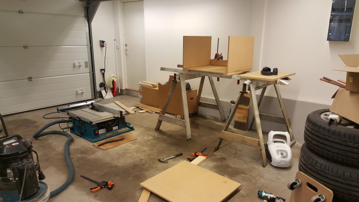 Byggprojekt i garage med bordssåg och improviserat rullbord på såghästar, verktyg och material på golvet.