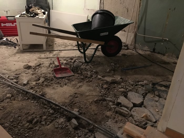 Påbörjad renovering med uppbilat betonggolv, skottkärra, hink och verktyg syns i ett rörigt utrymme.