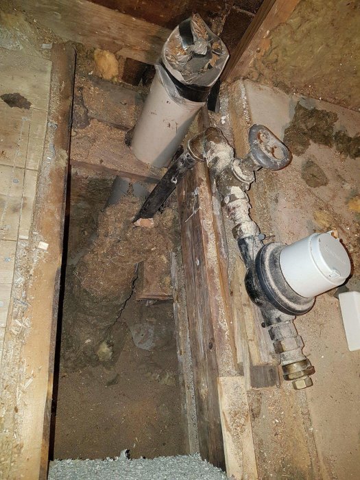 Vattenmätare och rör som kommer upp ur badrumsgolv med synlig bjälklag och isolering.