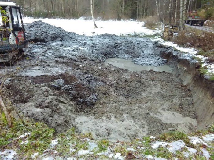 Utgrävning av en grop med jord och lera, bredvid grävmaskin, snötäckt mark i bakgrunden.